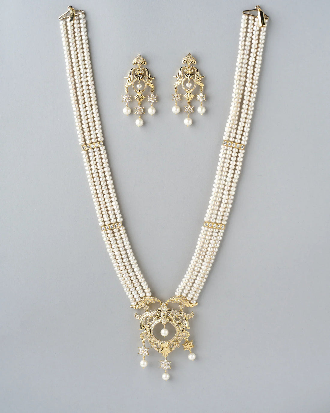 The Graceful Queen Pearl CZ Necklace Set by Chandrani Pearls features shimmering pearls, CZ work, a multi-strand necklace, and matching star-accented earrings with intricate designs displayed on brown fabric.