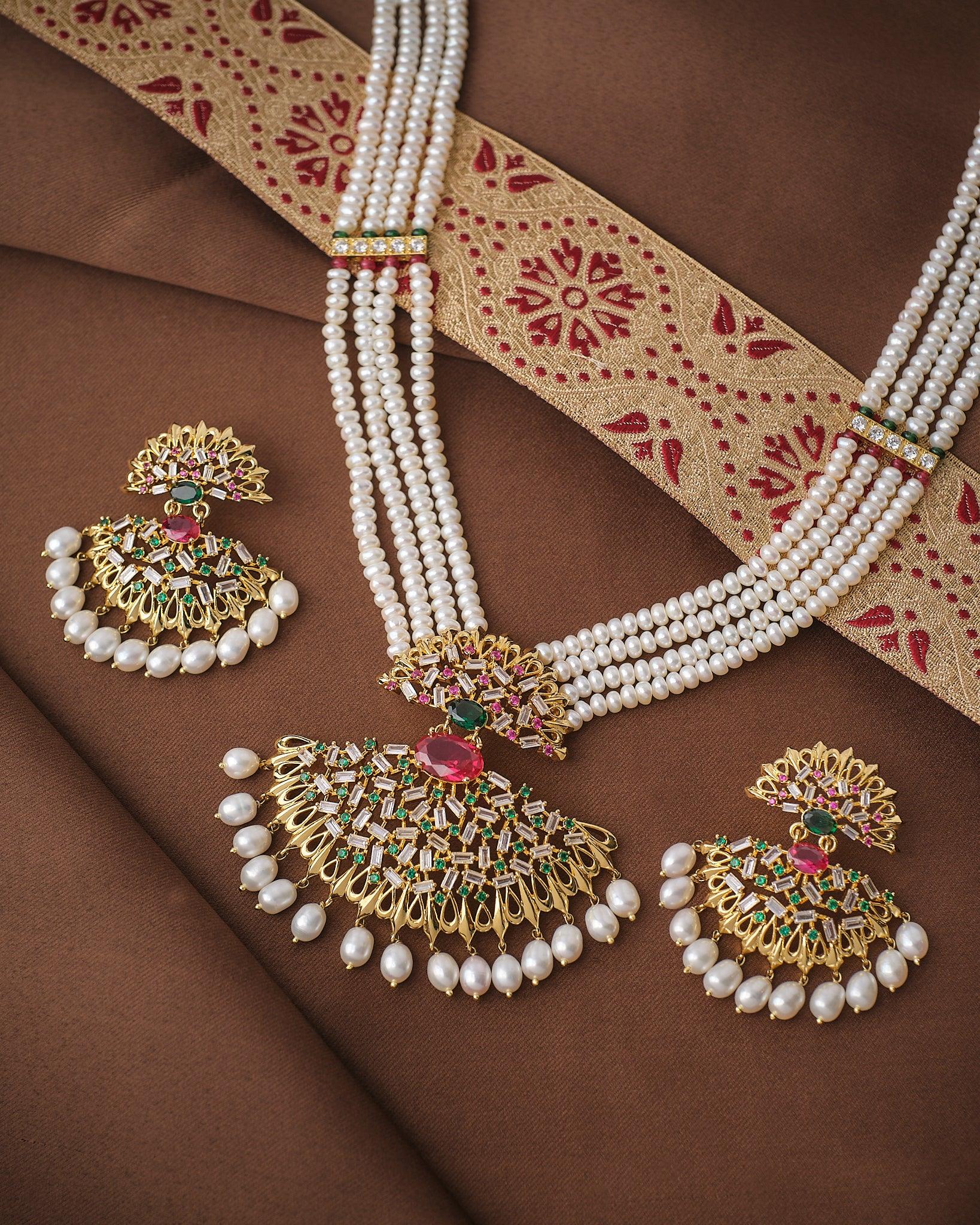 The Noor Queen’s Gaze Pearl CZ Necklace Set by Chandrani Pearls features a pearl-beaded necklace and matching earrings with CZ stones and gold, red, and green accents, elegantly displayed on brown fabric with a patterned border.