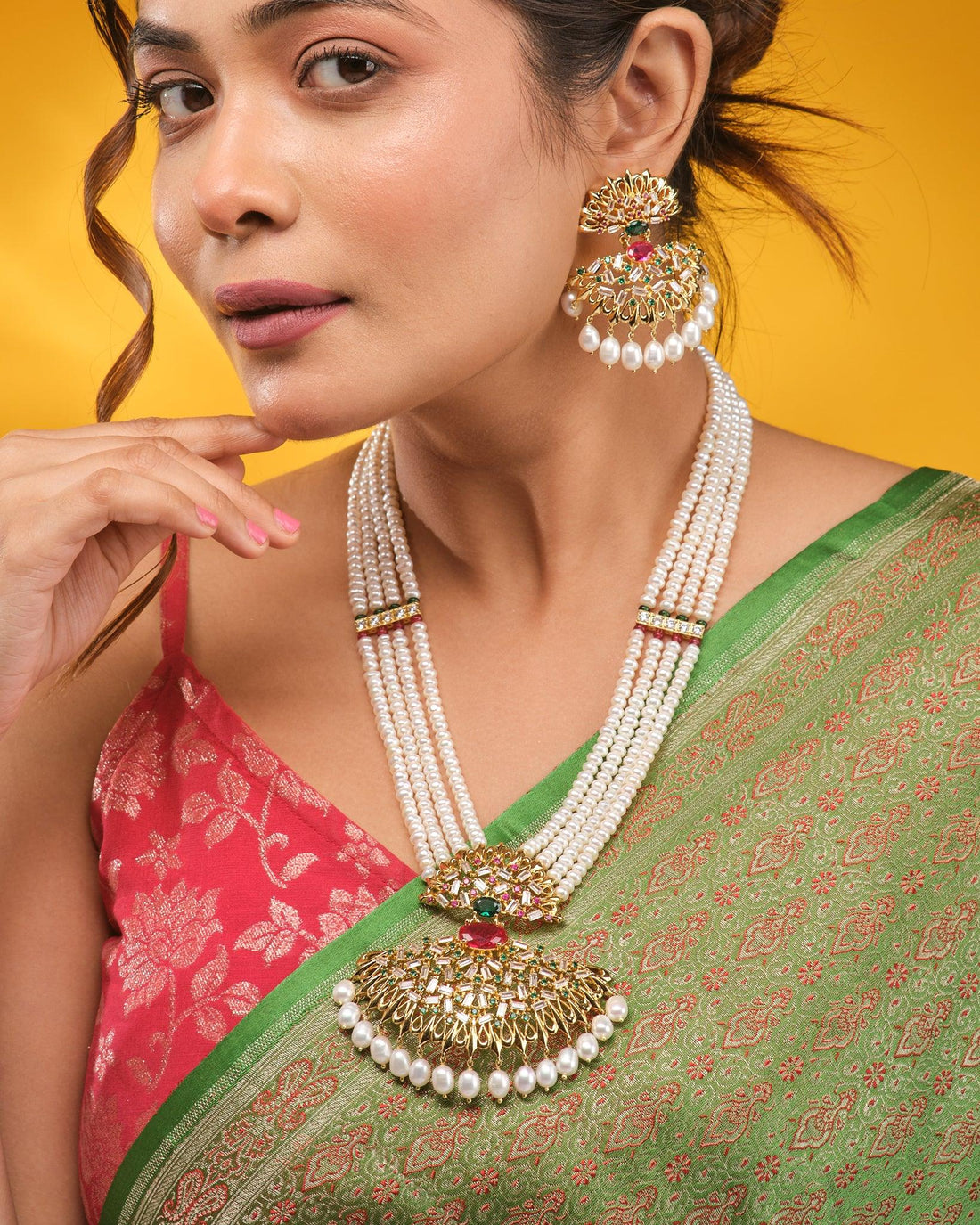 The Noor Queen’s Gaze Pearl CZ Necklace Set by Chandrani Pearls features a pearl-beaded necklace and matching earrings with CZ stones and gold, red, and green accents, elegantly displayed on brown fabric with a patterned border.