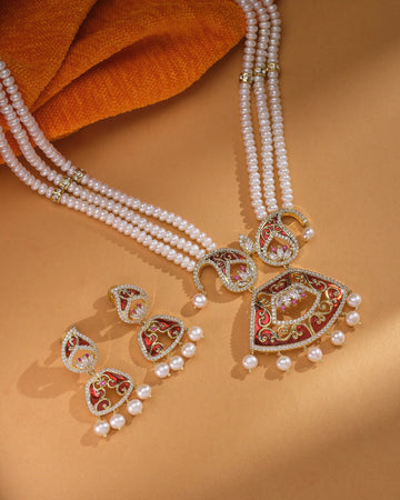 The Royal Sita Har Gorgeous Pearl Set by Chandrani Pearls features genuine pearls, gold-tone detailing, red and white enamel, and matching earrings—ideal for festive occasions. Shown on beige with orange fabric in the background.