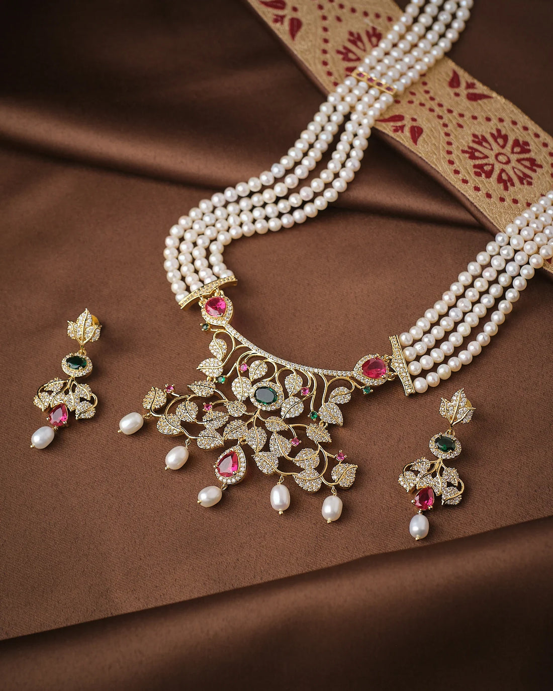 The Noor Floral Supreme Pearl CZ Sita Set by Chandrani Pearls features premium pearls, a gold and gemstone pendant with high-grade CZ stones, and matching earrings, beautifully displayed on brown fabric with a decorative border.