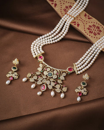 The Noor Floral Supreme Pearl CZ Sita Set by Chandrani Pearls features premium pearls, a gold and gemstone pendant with high-grade CZ stones, and matching earrings, beautifully displayed on brown fabric with a decorative border.