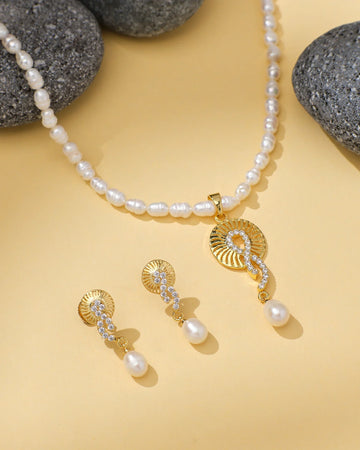 Chandrani Pearls' Eternal Grace Pearl Jewelry features a necklace with a gold and pearl pendant, matching gold earrings with pearl drops, and CZ accents, beautifully displayed on yellow with grey stones.
