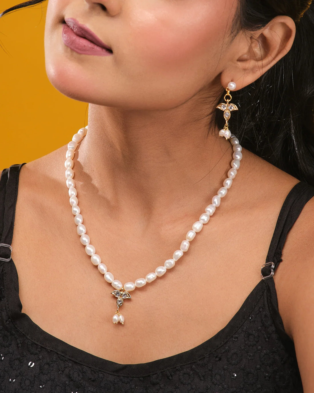 The Pearl Radiance CZ Necklace Set by Chandrani Pearls includes a radiant pendant and matching gold earrings with pearl and crystal details, beautifully arranged on a light yellow surface with black stones.