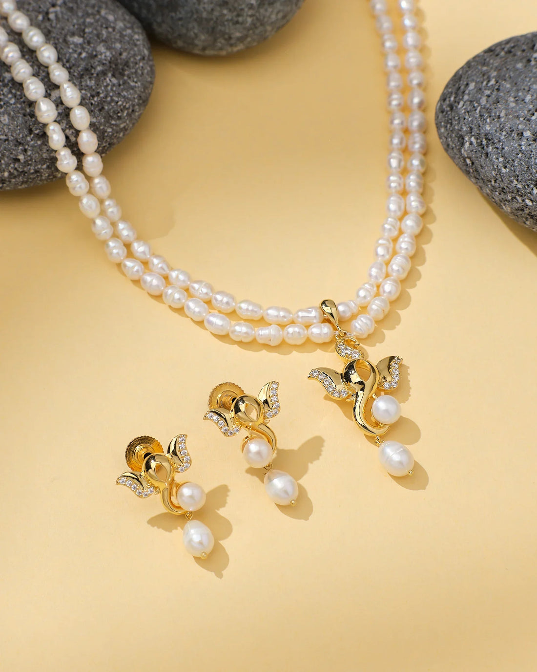 The Ganesh Inspired Pearl CZ Stone Set by Chandrani Pearls features double-strand pearls with a gold and pearl pendant, and matching gold, pearl, CZ stone & crystal earrings—perfect wedding jewelry shown on a yellow surface with gray stones.