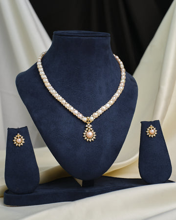 The Romantic Round CZ Necklace Set by Chandrani Pearls features a pearl necklace with a round CZ-accented pendant and matching earrings, elegantly displayed on a navy blue stand against a neutral draped backdrop.