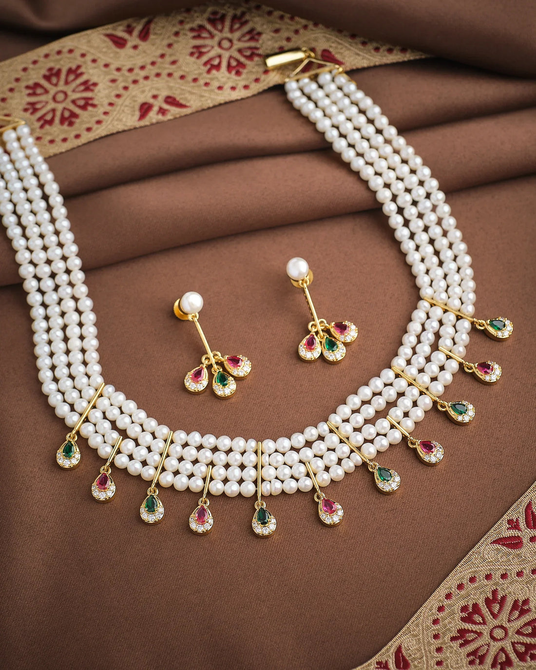 The Royal Whisper Pearl CZ Set by Chandrani Pearls features a three-strand pearl necklace with gold, green, and pink gemstone pendants, paired with matching pearl and gemstone drop earrings, shown on brown fabric.
