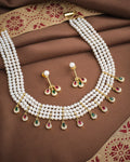 The Royal Whisper Pearl CZ Set by Chandrani Pearls features a three-strand pearl necklace with gold, green, and pink gemstone pendants, paired with matching pearl and gemstone drop earrings, shown on brown fabric.