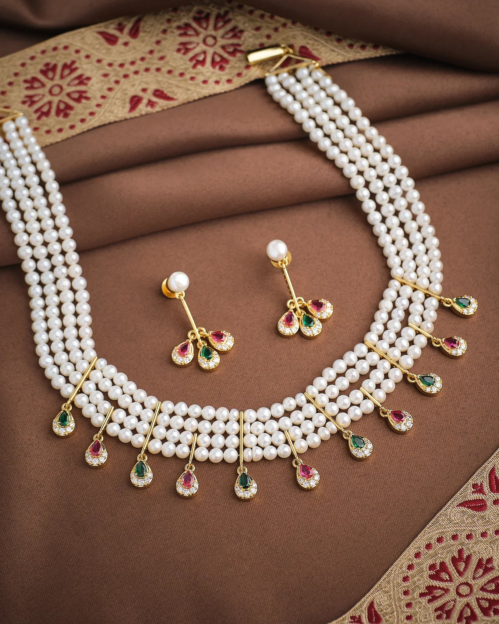 The Royal Whisper Pearl CZ Set by Chandrani Pearls features a three-strand pearl necklace with gold, green, and pink gemstone pendants, paired with matching pearl and gemstone drop earrings, shown on brown fabric.