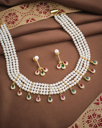 The Royal Whisper Pearl CZ Set by Chandrani Pearls features a three-strand pearl necklace with gold, green, and pink gemstone pendants, paired with matching pearl and gemstone drop earrings, shown on brown fabric.