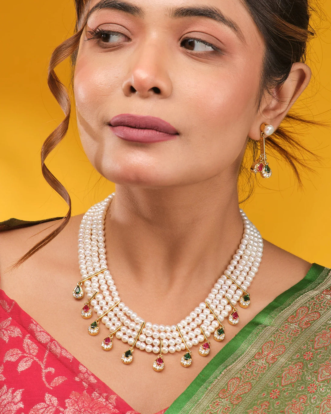 The Royal Whisper Pearl CZ Set by Chandrani Pearls features a three-strand pearl necklace with gold, green, and pink gemstone pendants, paired with matching pearl and gemstone drop earrings, shown on brown fabric.