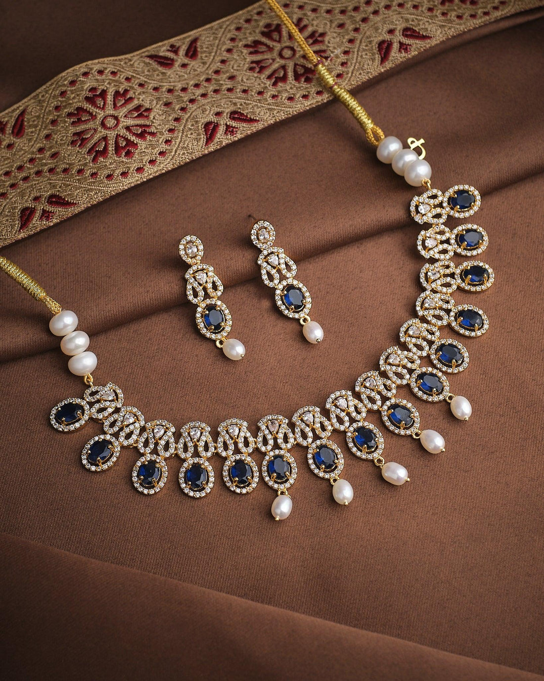 The Queen’s Radiant CZ Choker Set by Chandrani Pearls features a gold necklace and matching earrings adorned with blue stones, pearls, CZ stones, and clear crystals, displayed on luxurious brown and gold embroidered fabric.