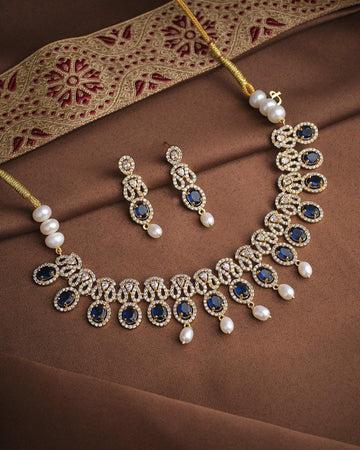 The Queen’s Radiant CZ Choker Set by Chandrani Pearls features a gold necklace and matching earrings adorned with blue stones, pearls, CZ stones, and clear crystals, displayed on luxurious brown and gold embroidered fabric.
