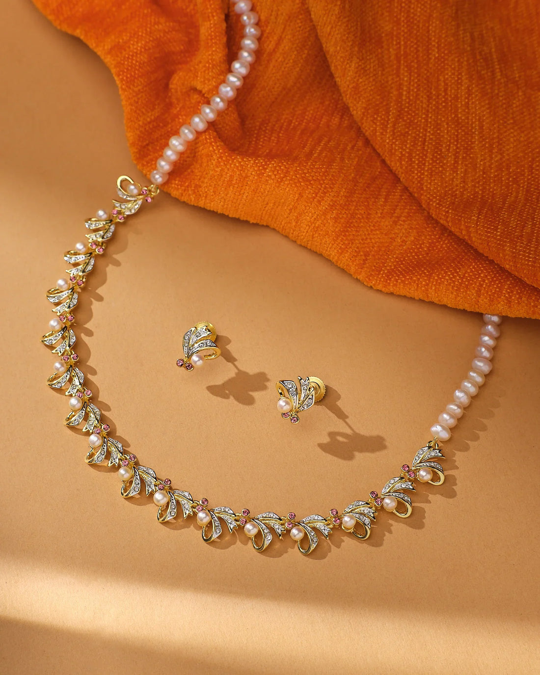 The Twilight Pearl Radiance Set by Chandrani Pearls—a gold and natural pearls necklace with matching stud earrings—is displayed on a tan surface beside orange fabric, perfect for occasion wear.