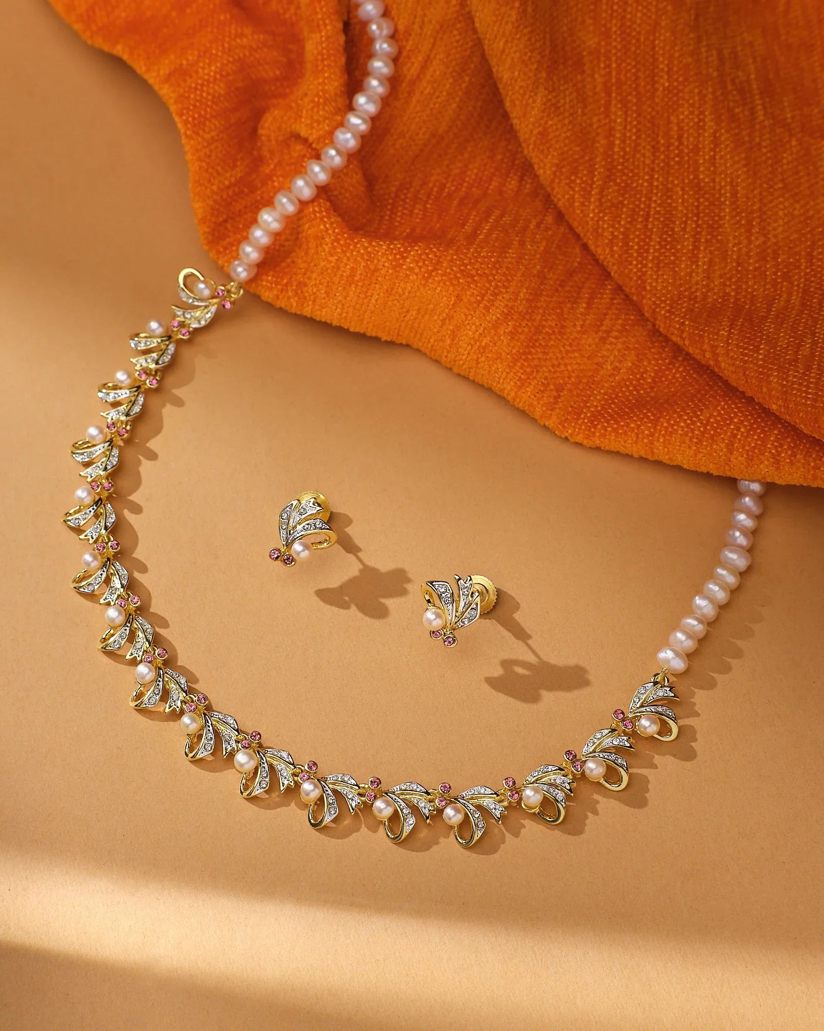 The Twilight Pearl Radiance Set by Chandrani Pearls—a gold and natural pearls necklace with matching stud earrings—is displayed on a tan surface beside orange fabric, perfect for occasion wear.