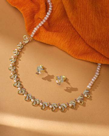 The Twilight Pearl Radiance Set by Chandrani Pearls—a gold and natural pearls necklace with matching stud earrings—is displayed on a tan surface beside orange fabric, perfect for occasion wear.
