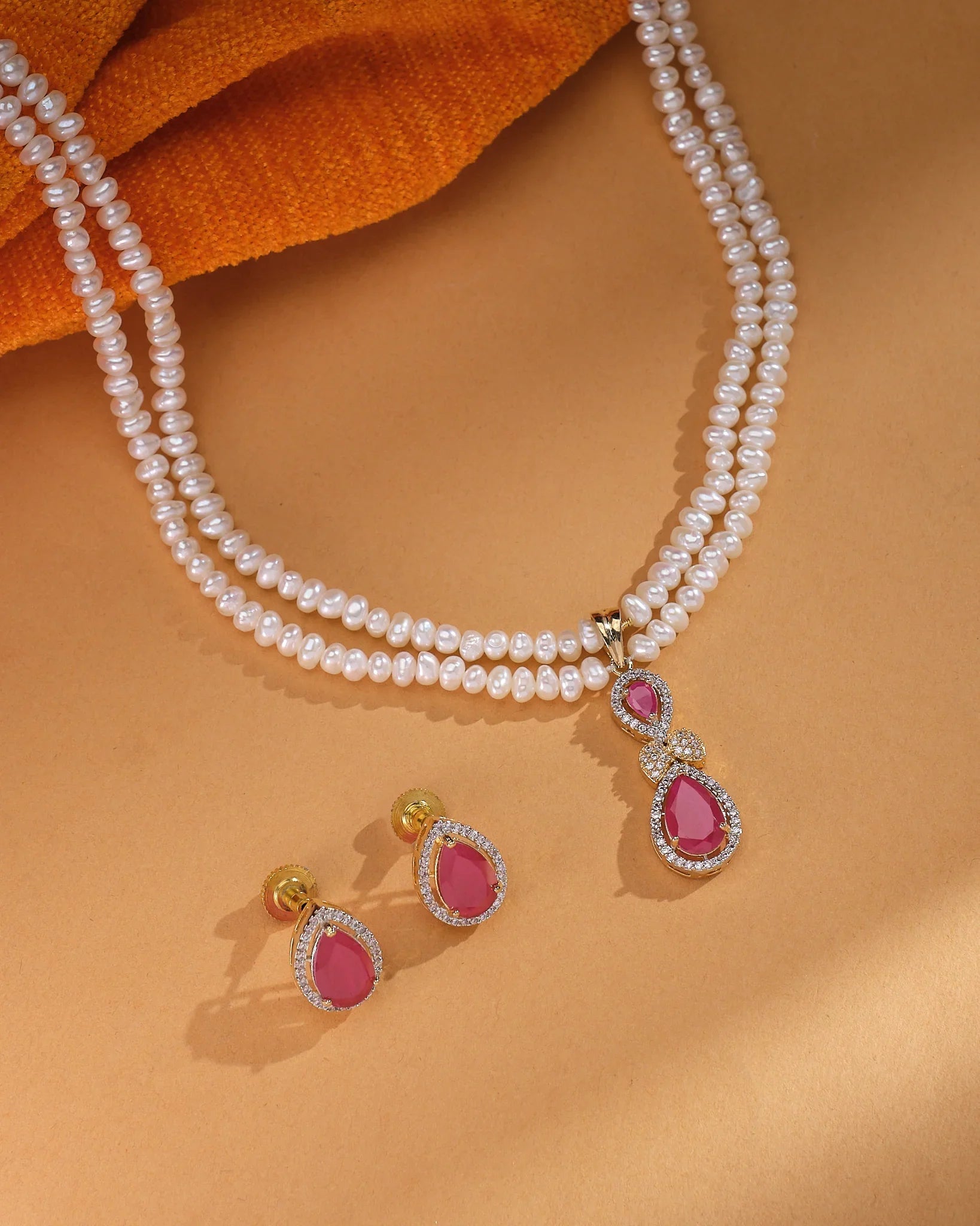 The Minimalist Real CZ Pearl Set by Chandrani Pearls features a double-stranded pearl necklace with a ruby and diamond pendant and matching earrings, displayed on a tan surface with an orange cloth—ideal for lovers of elegant minimal jewelry.