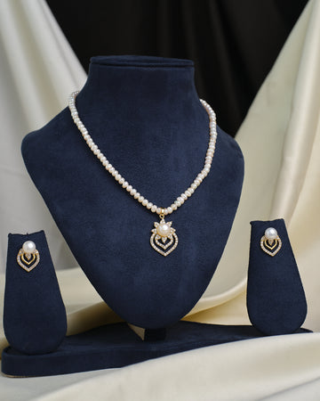 The Chandrani Pearls Elegant Teardrop CZ Set features a pearl necklace with a gold and pearl pendant, displayed on a navy blue stand with matching teardrop earrings, set against draped fabric.