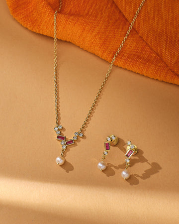 The Pure Elegance Pretty Set by Chandrani Pearls features a gold necklace and matching earrings with geometric designs, red accents, CZ stones, and pearl drops—ideal for formal occasions or gifting—shown on beige with orange textured fabric.