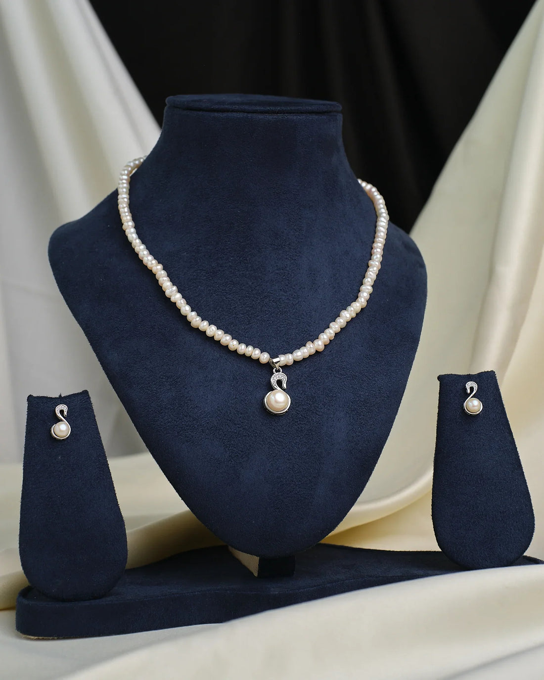 The Chandrani Pearls CZ Halo Pendant Set features a pearl necklace with matching pendant and earrings, each accented by CZ stone halos, elegantly displayed on navy blue velvet against a cream and black fabric background.