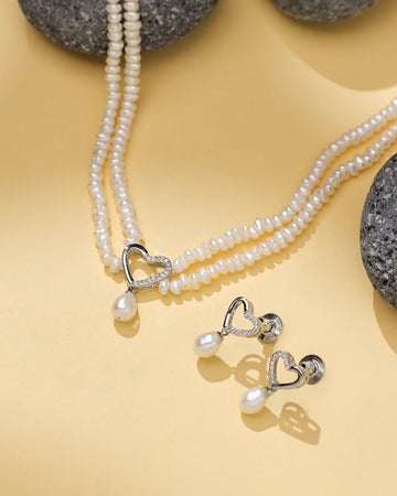 The Timeless Pearl Treasure Set by Chandrani Pearls, featuring two pearl necklaces and matching heart-accented earrings, is elegantly displayed on a beige surface with gray stones, highlighting the classic charm of pearls.