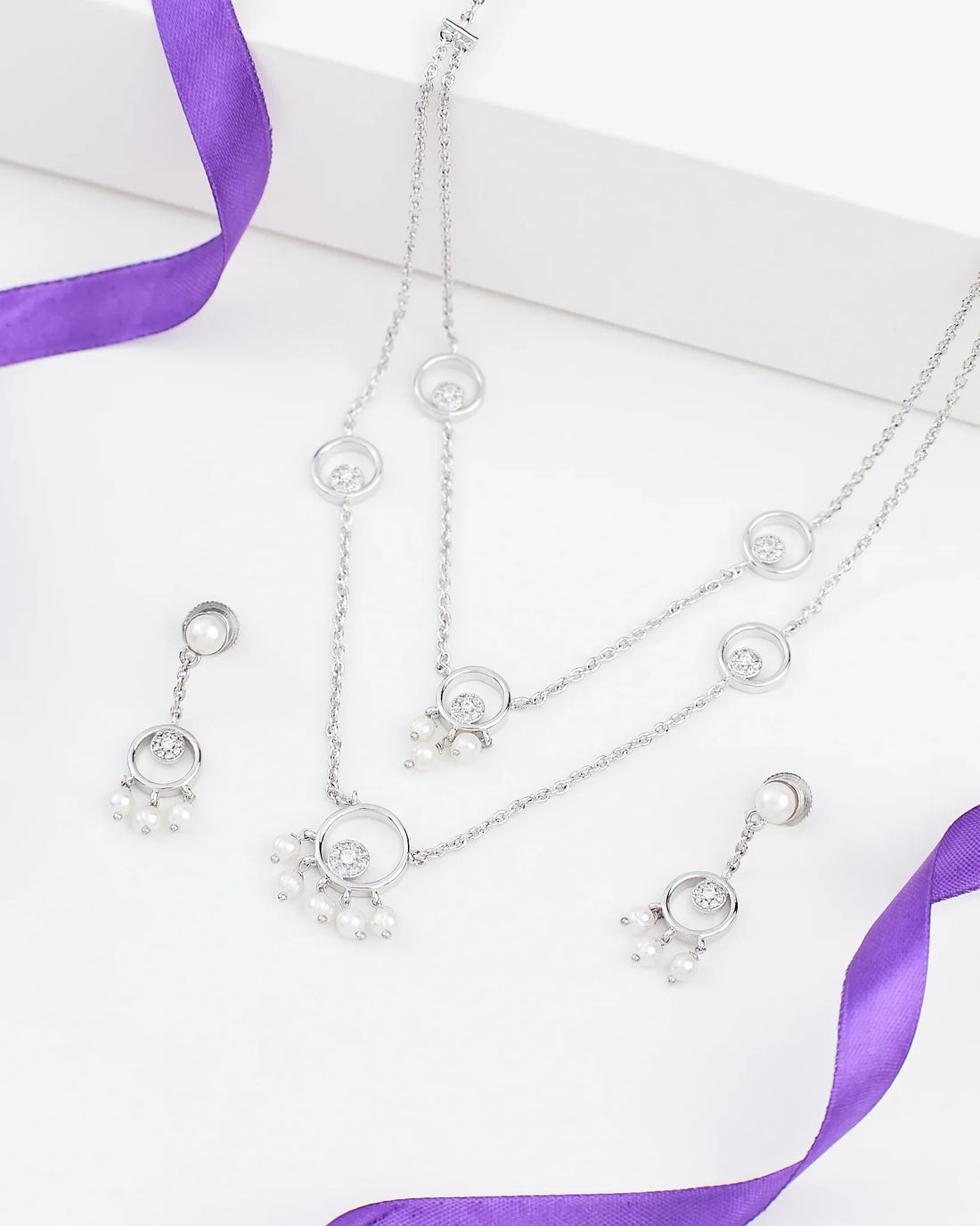The Demifine CZ Necklace Set Graceful & Glamorous by Chandrani Pearls features circular pendants, pearl accents, and sparkling CZ stones, elegantly displayed on a white surface with a purple ribbon.