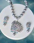 The Chandrani Pearls Dainty Pearl & CZ Jewelry Set includes a gray beaded necklace with an ornate diamond, CZ stones, and blue gemstone pendant, plus matching earrings, displayed on a round white surface.
