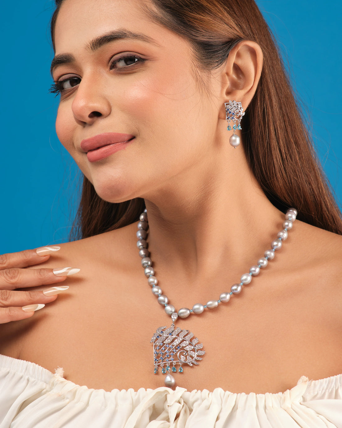 The Chandrani Pearls Dainty Pearl & CZ Jewelry Set includes a gray beaded necklace with an ornate diamond, CZ stones, and blue gemstone pendant, plus matching earrings, displayed on a round white surface.