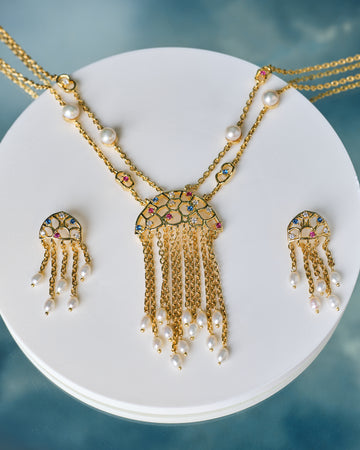 The Trendy Handcrafted Chain & CZ Necklace Set by Chandrani Pearls features colorful gemstones, CZ stones, and pearls in a multi-chain design with matching earrings, elegantly displayed on a white circular stand.