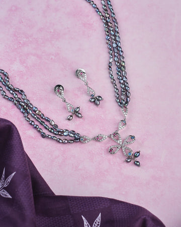 The Florial Pearl Black Set by Chandrani Pearls features a dark pearl necklace and matching earrings, elegantly displayed on a light pink surface next to folded purple fabric.