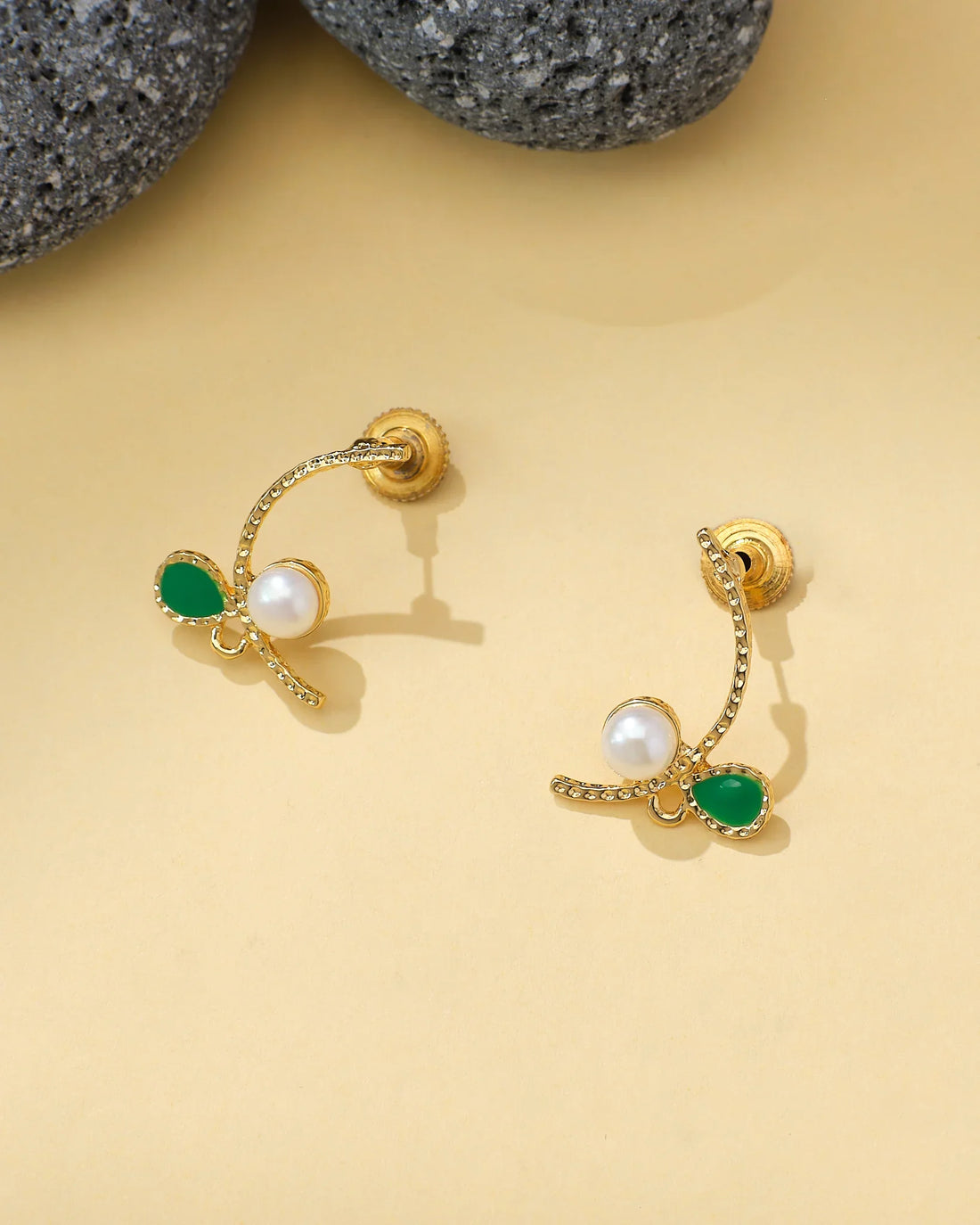 The Halo CZ & Pearl Stud Earrings Half Classic Brilliance by Chandrani Pearls, gold-toned with green teardrop gemstones, CZ stones, and central pearls, are displayed on a light yellow surface near gray stones.