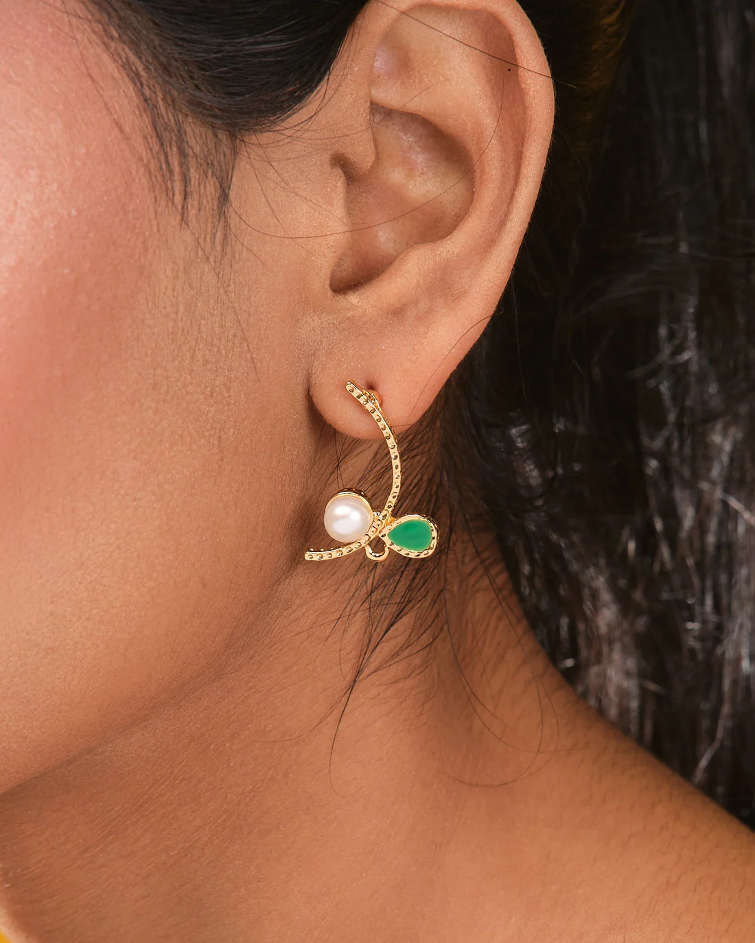 The Halo CZ & Pearl Stud Earrings Half Classic Brilliance by Chandrani Pearls, gold-toned with green teardrop gemstones, CZ stones, and central pearls, are displayed on a light yellow surface near gray stones.