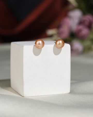 Simple Stud Earrings by Chandrani Pearls India, featuring round peach-colored pearls, are showcased on a white block with a blurred floral background, highlighting their elegant and sophisticated design.
