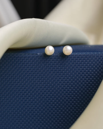 Two Simple stud white pearl earrings from Chandrani Pearls are displayed on a textured blue surface with soft white fabric, adding timeless charm to your collection.