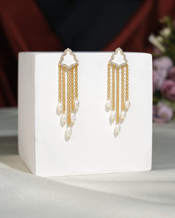 The Stylish Hang Earrings by Chandrani Pearls India, featuring gold and pearls, are displayed on a white cube with a blurred floral background.
