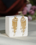 Fashionable CZ Hang Earrings by Chandrani Pearls India, featuring gold finish, lustrous pearls, and chain details, displayed on a white block with a blurred floral background.