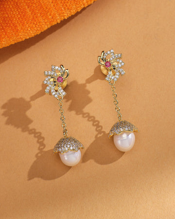 The Pearl Radiance CZ Jhumka by Chandrani Pearls features gold drop earrings with floral diamond-style tops, chain links, and round pearls in pave-set caps—festive jewelry elegantly displayed on a tan surface with an orange fabric accent.