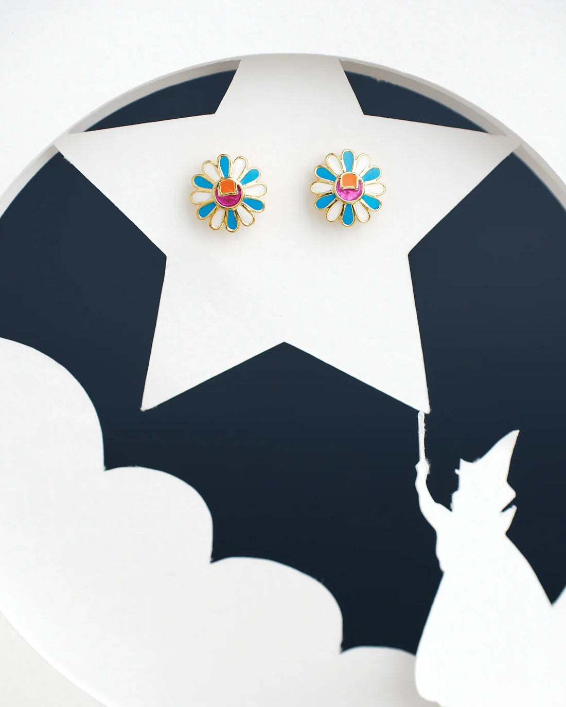 Chandrani Pearls India's Sunflower Stud Earrings, featuring two daisy-shaped designs with blue petals and red centers, are displayed on a circular background with a silhouette of a person pointing to a star.