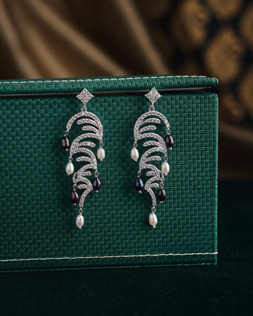 A pair of ornate earrings from Chandrani Pearls, named The Ratriratna-Symbolizing Purity, featuring silver design with pearls and black beads, are displayed on a textured green surface.