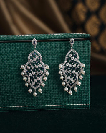 Navodha - The Wedded Dazzle Pearl Earrings by Chandrani Pearls, featuring diamond patterns and pearl accents, elegantly displayed on a textured green surface.