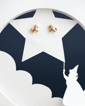 A pair of The Cute Foal Stud Earrings by Chandrani Pearls India with pink manes sits on a white star cutout against a dark background, beneath which a wizard silhouette points a wand.