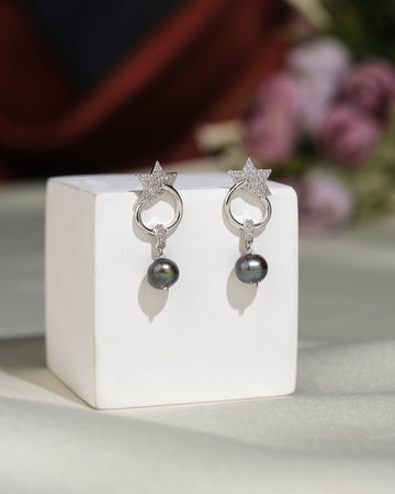 The Trendy CZ Hang Earrings by Chandrani Pearls India feature star-shaped silver designs with black pearls, displayed on a white stand against a blurred floral background—perfect for adding sophistication to any look.