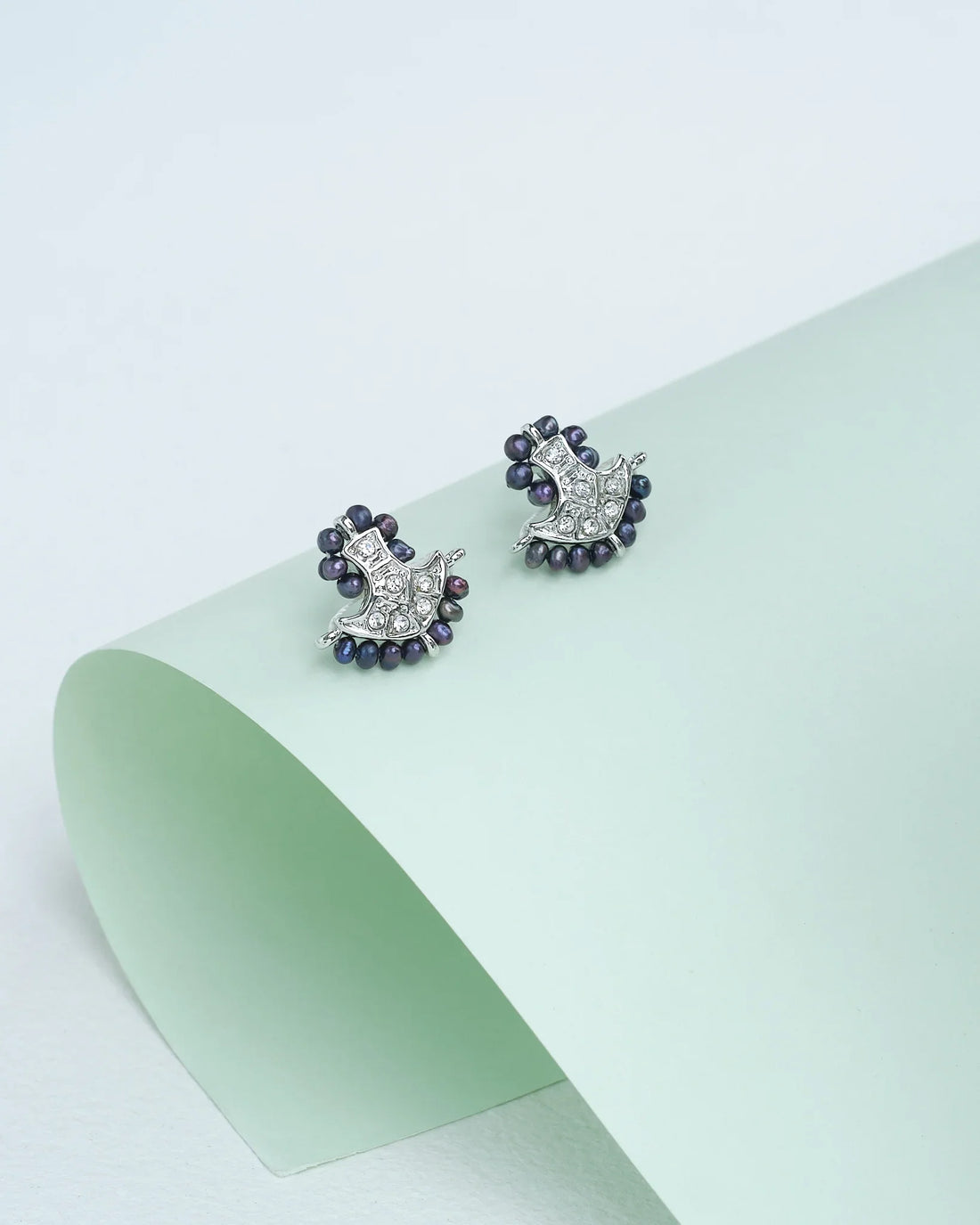 A pair of "Lustrous pearls, radiant CZ" earrings by Chandrani Pearls, featuring small dark beads and clear stones, displayed on a curved pale green surface.