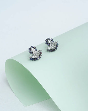 A pair of "Lustrous pearls, radiant CZ" earrings by Chandrani Pearls, featuring small dark beads and clear stones, displayed on a curved pale green surface.