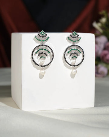 The Fashionable CZ Hang Earrings by Chandrani Pearls India feature green and black stones, circular designs, and elegant pearl drops, displayed on a white cube with a blurred floral background for timeless appeal.