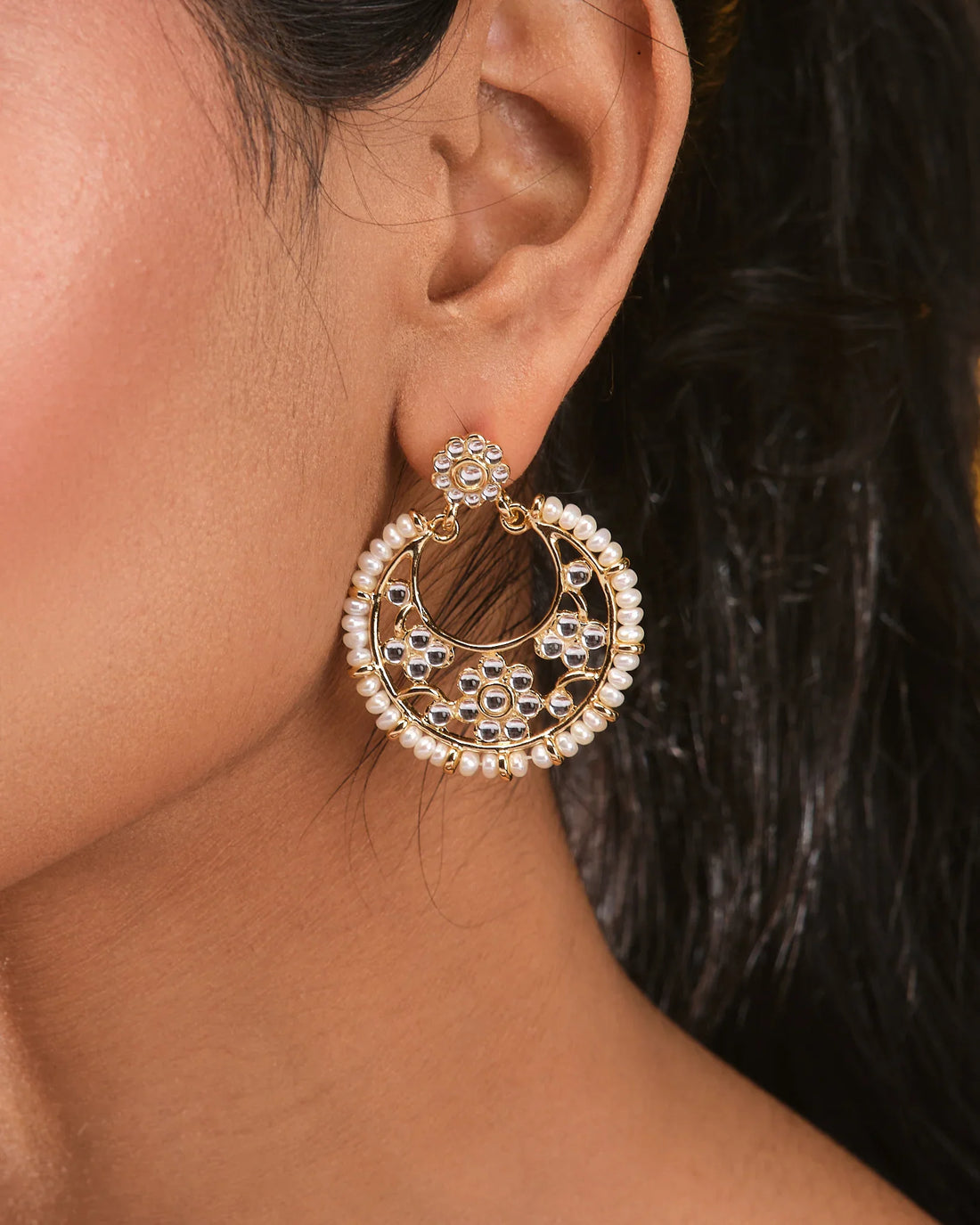 The Gold Plated Pearl Earrings with CZ Flowers by Chandrani Pearls feature feminine gold-tone hoops with floral CZ motifs and pearl accents, making them perfect for lovers of elegant jewelry. Displayed on beige with gray stones in the background.