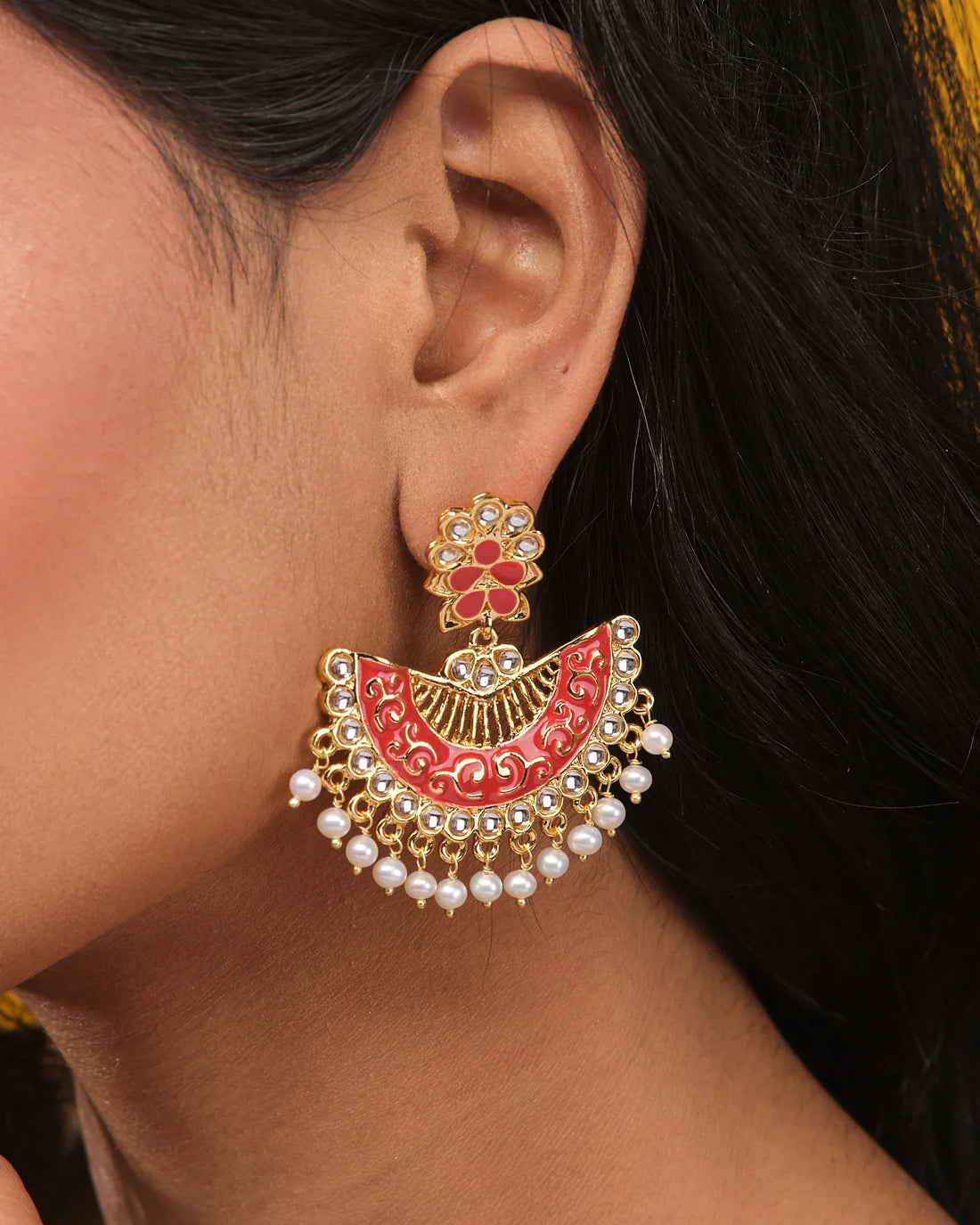 The Chandrani Pearls Festive Shine Enamel Pearl Jhumka features gold-toned and red enamel floral designs with CZ accents and dangling white pearls, presented on a beige surface.