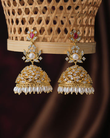 The Noor Celestial Pearl Jhumkas by Chandrani Pearls feature ornate gold, white stones, floral motifs, and delicate pearl drops, creating an elegant and timeless look against a woven basket background.