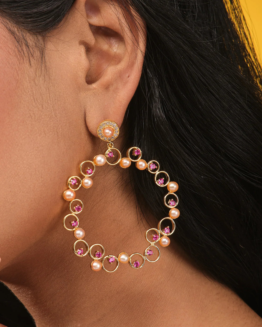 The Timeless Elegance Pearl CZ Jhumka by Chandrani Pearls features large circular gold jhumkas with pink gemstones, CZ stones, and natural pearls, displayed on a light brown surface with orange fabric in the corner.
