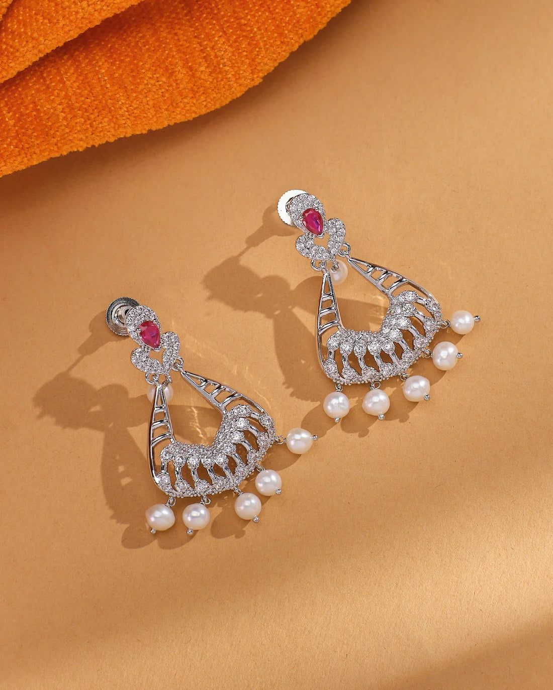 The Regal Pearl Cascade Jhumka by Chandrani Pearls features silver CZ work, intricate designs, pink stones at the top, and pearl drops—ideal wedding jewelry—shown on a tan surface with orange fabric in the background.